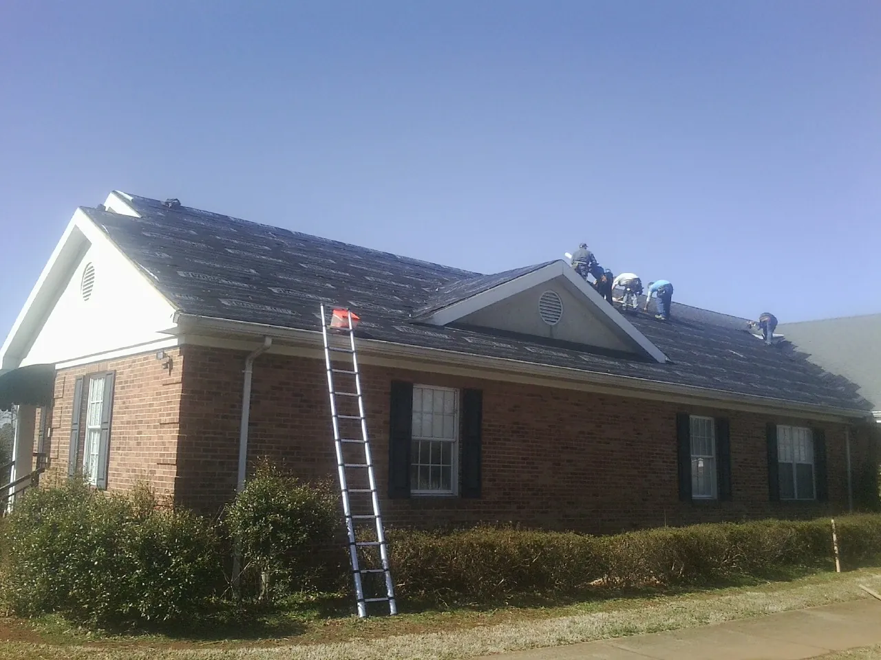 Crew tearing shingles off a brick ranch roof with a ladder set — Metal Roof Repair work by our crew in Edgerton